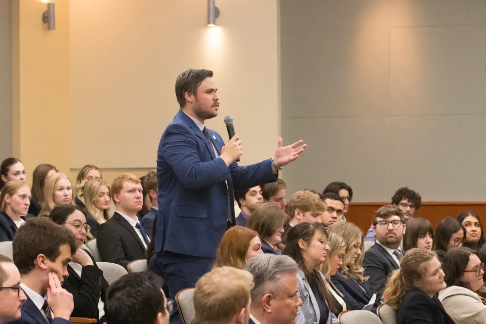A student stands up in a crowd and speaks into a microphone.
